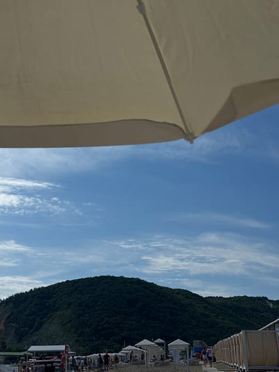 The image depicts a beach scene on a sunny day with a large, light-colored umbrella dominating the foreground. In the background, a lush green, tree-covered mountain slopes down towards the sea. The sky is blue with scattered white clouds. Below the mountain, the beach is populated with people, beach umbrellas, and structures that appear to be changing rooms or cabanas. The presence of these amenities suggests this is a developed recreational area. The overall impression is one of a typical summer day at a coastal resort.