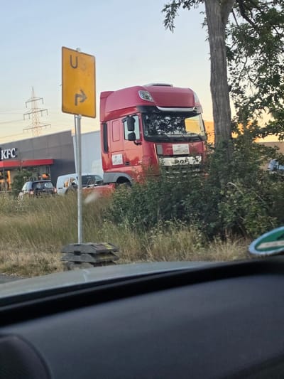 The image shows a red DAF semi-truck parked on a grassy area near a KFC restaurant. A yellow directional sign with "U3" and a right arrow is visible to the left of the truck. In the background, a large electrical pylon and a building with a KFC sign are present. Several other vehicles, including a white van and a dark-colored car, are parked in front of the KFC. The scene appears to be a roadside area, possibly a rest stop or a parking lot adjacent to the restaurant. The overall lighting suggests it is either early morning or late afternoon.