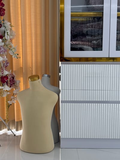 In Tanjungmeru, Indonesia, a mannequin torso, a light beige colored, is positioned in front of a golden draped curtain. Behind it, another mannequin torso, this one grey, is visible. To the right, a white cabinet with vertical grooves and a gold trim is displayed. The cabinet has glass doors revealing stacks of clothing. The floor is tiled in a light color. A floral arrangement with white, pink, and burgundy flowers hangs to the left of the mannequins.