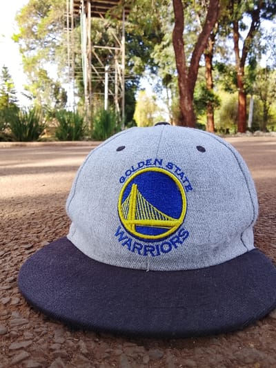 A gray baseball cap with a dark navy blue brim rests on a reddish-brown, textured ground surface, possibly compacted dirt or gravel. The cap's front displays an embroidered Golden State Warriors logo: a yellow outline of the Golden Gate Bridge within a blue circle, with "GOLDEN STATE" arced above and "WARRIORS" arced below in blue text. In the background, a wider, lighter-colored pathway is visible, flanked by green shrubs and trees with reddish-brown trunks. A tall, multi-level metallic lattice structure, consistent with a water or communication tower, stands prominently on the left side of the background. The scene is illuminated by natural daylight. No individuals, actions, or direct interactions are depicted. This scene is located in Kikuyu Township ward, Kenya.