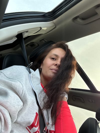 A woman with long, wavy dark brown hair is captured in a close-up, low-angle shot from inside a car, presumably in Minderevo, Russia. She is seated in the passenger seat, wearing a light grey zipped hoodie with red accents and a red long-sleeved top underneath. A black seatbelt is visible across her chest. She looks directly at the camera with a neutral expression. The car's interior is visible around her, including parts of the light-colored headliner, a grab handle, the A-pillar, and a black leather seat with white stitching. Above her, a bright, overcast sky is visible through the car's windows and possibly a sunroof, indicating it is daytime. There is partially obscured red text on her hoodie that is not fully decipherable. The overall scene suggests she is traveling or taking a break inside a vehicle.