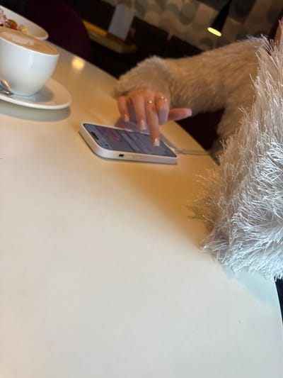 A close-up indoor shot depicts a light beige table with a smartphone, a cup of coffee, and a hand with rings on it. The fingers are tapping on the screen of the smartphone. The smartphone has a white case. The coffee cup appears to be white and partially obscures a saucer and a bowl of food. A fuzzy white garment is visible in the top left of the frame. The location and city cannot be determined from the image.