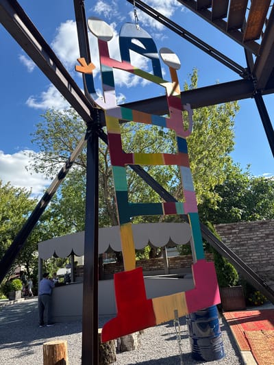 A bright, colorful, abstract metal sculpture resembling a ladder or abstract figure is suspended from a dark metal framework against a bright blue sky with white clouds. The sculpture is composed of various geometric shapes painted in a rainbow of colors, including red, orange, yellow, green, teal, light blue, pink, and purple. It appears to be an outdoor installation.
In the background, a man wearing a blue plaid shirt and gray pants is standing at a gray kiosk with a scalloped awning. Two people are visible behind the counter of the kiosk. To the right of the sculpture, a portion of a red patterned rug is visible on the ground, and a dark blue barrel is positioned beneath the lower part of the sculpture. The ground is covered in small gray gravel. Trees with green foliage are visible behind the sculpture and the kiosk. The overall environment suggests an outdoor public space. The location is identified as Yelets, Russia.