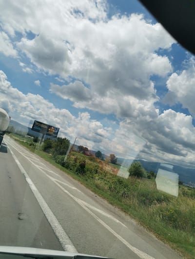 The image captures a scenic view from a vehicle moving along a highway, likely in or near the city of Krivina, Bulgaria. The foreground shows a paved road with white lane markings, indicating a clear path for traffic. To the right of the road, a lush green embankment covered with tall grass and scattered wildflowers slopes upwards. In the background, a billboard advertising a casino with the text "Оферти и печалби като в света" and the number "8888" is visible. Beyond the billboard, a line of trees forms a natural barrier, with rolling hills and distant mountains completing the landscape. The sky is a dynamic canvas of blue, dotted with large, fluffy white clouds, suggesting fair weather with some cloud cover. Reflections on the vehicle's window are also present, obscuring parts of the scene.