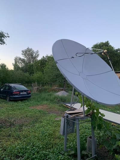 This media file captures an outdoor scene dominated by a large, pale satellite dish mounted on a metal stand. The dish is positioned prominently in the foreground, angled upwards towards a clear, pale blue sky. Lush green foliage, including trees and dense ground cover, surrounds the satellite dish and extends into the background. A dark-colored car is parked amidst the greenery on the right side of the image.
The overall impression is one of a rural or semi-rural setting, possibly a backyard or a less manicured area. The lighting suggests it is either early morning or late afternoon due to the soft, diffused light. There are no people visible in the frame.
Notable details include the construction of the satellite dish mount, which appears to be made of weathered metal. Vines and leaves from a nearby plant are encroaching on the metal structure. On the car's license plate, the visible text is "980BEM 13". The location context provided, Зертас ауылдық округі, Kazakhstan, indicates the geographical setting. The scene appears tranquil, with no discernible activity taking place beyond the static presence of the objects and vegetation. The weather seems calm and clear.