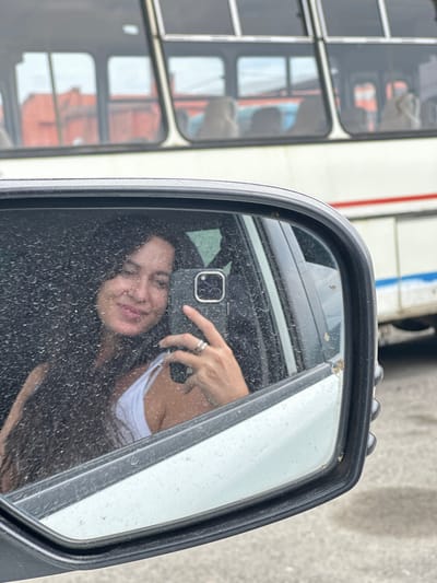 A woman with long dark hair is reflected in a car's side mirror, taking a selfie. She is wearing a white tank top and rings on her fingers. The mirror's surface is covered in water droplets, creating a distorted view of her face. In the background, a white bus with red and blue stripes is visible, its windows reflecting an orange building. The image appears to be taken in Slyudyanka, Russia, though this cannot be definitively confirmed from the visual evidence alone.