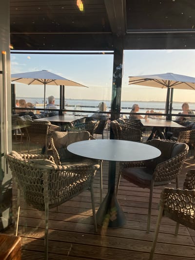The image depicts an outdoor seating area of a restaurant or cafe in Hüde, Germany, likely overlooking the artificial lake known as Dümmer See. The time of day appears to be late afternoon or early evening, indicated by the warm, low-angle sunlight casting long shadows across the wooden deck floor.
In the foreground, a round, dark-colored table with a tapered base is positioned. Surrounding this table are several chairs, all featuring a woven, wicker-like design with thick cushions. One chair in the immediate foreground is angled towards the viewer, showcasing its detailed construction.
In the middle ground, there are more tables and chairs, some occupied by diners. Through the glass enclosure of the restaurant, several people are visible seated at tables. One man with grey hair is facing away from the camera, seated at a rectangular table. To his right, another person is seated. Further to the right, two women are seated at a table, with one looking out towards the water. Beyond these tables, white umbrellas are deployed, providing shade.
The background features a body of water, presumably Dümmer See, with a clear sky. The water is calm, with a few distant figures visible near a railing along the shore, suggesting a promenade or pier. The overall scene conveys a relaxed atmosphere, typical of a lakeside dining establishment. The presence of what appear to be wind turbines in the far distance suggests the location is in a relatively open, possibly rural area surrounding the lake.
