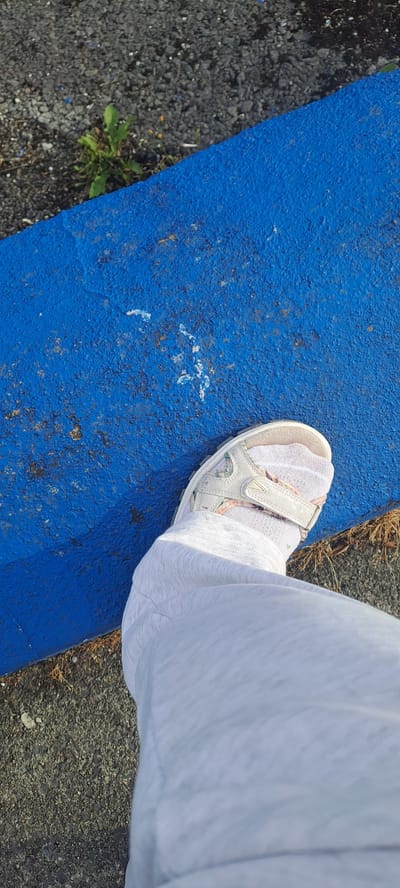 An overhead shot shows a left foot in a white sandal with a floral pattern and a white sock, stepping onto a textured blue surface. The foot is clad in light grey pants. The blue surface appears to be painted asphalt, possibly a speed bump or curb, with some scuff marks and debris. To the left of the blue surface is dark grey asphalt with scattered small stones and a small patch of green weeds. To the right of the blue surface is more dark grey asphalt with dry, brown grass-like material. The lighting suggests it is daytime, with shadows cast on the blue surface. The image's perspective is from above, looking down at the foot and the ground. This scene appears to be an outdoor public space, perhaps a park, playground, or street in Castlebar, Ireland.