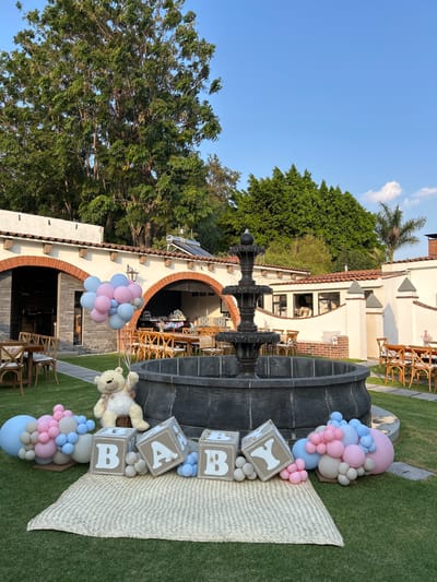 The image shows a baby shower setup in what appears to be an outdoor garden setting. It is likely in Santa Cruz Tehuixpango, Mexico, based on the information provided. A tiered fountain is the main focal point, surrounded by a grassy area. The decorations include large "BABY" blocks, arrangements of pastel pink and blue balloons, and a teddy bear. A woven mat is placed on the grass. In the background, a building with an arched entryway is visible, with tables and chairs set up inside, suggesting a reception area. Other buildings and trees are present in the background under a clear, blue sky.