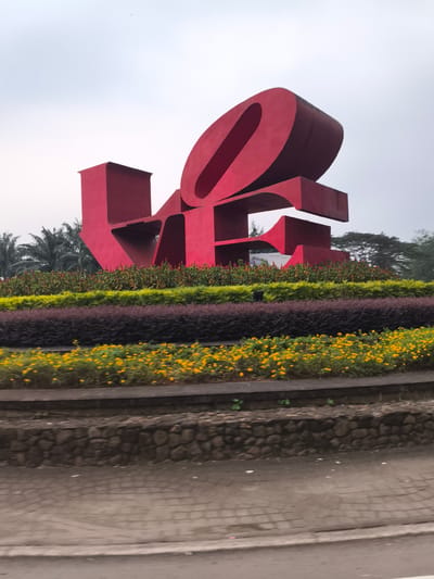 The image displays a large, red, three-dimensional sculpture spelling out the word "LOVE" under an overcast sky in Babakan Madang, Indonesia. The prominent sculpture is the central subject, set within a meticulously designed garden.
In the foreground, a paved surface, possibly a sidewalk or road, transitions into a low, rugged stone retaining wall. Above the wall, the garden features several distinct layers of plants and flowers. The outermost layer consists of bright yellow flowers, followed by a dense row of dark reddish-purple shrubs. Behind these, a band of lighter green foliage provides contrast, while plants with reddish-orange flowers or berries are situated closest to the sculpture's base.
Behind the landscaping, tropical palm trees are visible on the left, and other green trees stretch into the distance on the right. The sky is a uniform, light gray, indicating a cloudy or hazy daytime conditions, with no direct sunlight. No people are present, and no particular activity is taking place; the scene is static, showcasing a public art installation and its surrounding greenery. The text "LOVE" is clearly visible, forming the entirety of the bold red sculpture.