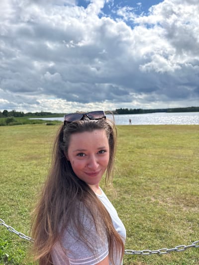The image features a young woman posing for a selfie outdoors. She has long, brown hair and is wearing sunglasses on top of her head. She is looking at the camera with a relaxed expression. The background shows a body of water with a faint reflection of the sky, suggesting it is a sunny day. To the right of the water, there is a grassy field. In the distance, a lone figure can be seen near the water's edge. The sky is partly cloudy with dramatic cloud formations. A metal chain, likely part of a swing set, is visible in the lower right corner of the frame. The orientation of the image suggests it might have been taken in a vertical format. The scene appears to be a natural, possibly recreational area, consistent with the provided location context of Мазалаўскі сельскі Савет, Belarus.