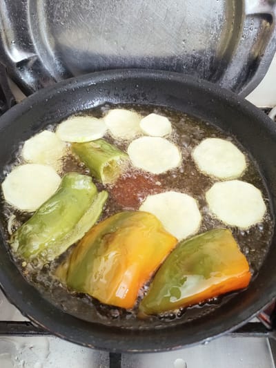 The image shows a close-up, overhead view of a frying pan filled with hot oil, in which several pieces of food are being cooked. The pan is black and has a metallic handle. In the pan, there are sliced rounds of what appear to be potatoes, scattered amongst pieces of bell peppers. The bell peppers are cut into large chunks, with some being green and others a mix of green and orange. The oil is bubbling vigorously around the food, indicating it is hot. The overall scene suggests food preparation. The background shows a reflective, scratched metallic surface, possibly a stovetop or a lid, with water droplets visible on it. There are no people visible in the frame. The lighting suggests it is daytime, and the context of a kitchen setting is implied by the presence of the frying pan and stovetop. No visible text is present in the image. The location context of Ariana, Tunisia, suggests this is likely a dish being prepared in a Tunisian household.