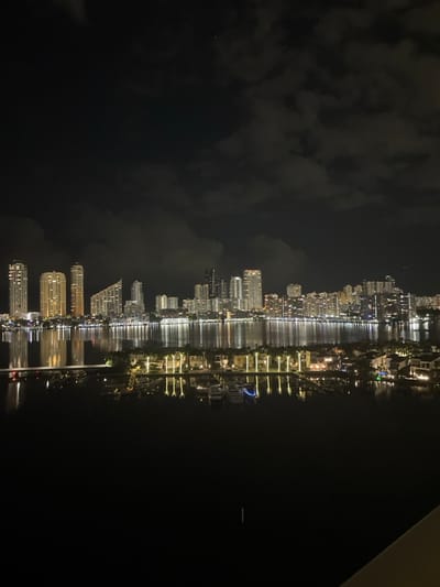 Nighttime view of a cityscape reflected in water. Tall buildings, illuminated with lights, line the waterfront. The reflection mirrors the city, creating a doubled visual effect. Dark clouds fill the sky above the city. The overall scene evokes a sense of urban beauty at night, with the reflections adding depth and complexity.