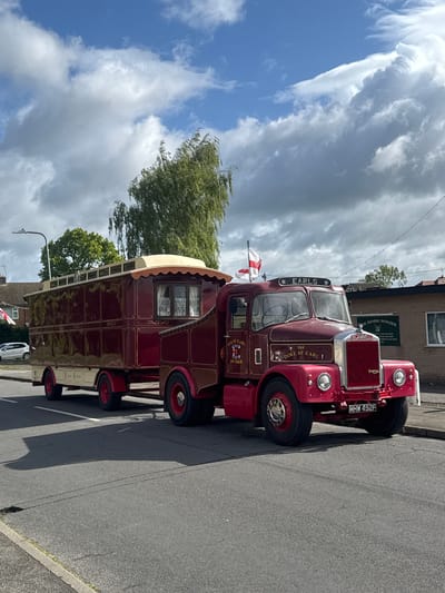 The image captures a maroon and cream Scammell Showtrac, bearing the name "EARLS THE DUKE OF EARL," parked on a street in South Kesteven, United Kingdom. The vehicle, registered as MHW 492F, includes a trailer-like structure attached. The sky is partly cloudy. A brick building is partially visible in the background. A street lamp is visible on the left side of the image. The scene appears static, with no visible people or ongoing actions.