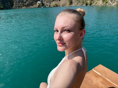 The image depicts a young woman with blonde hair pulled into a bun, wearing a white crocheted bikini top. She is seated on a wooden dock, with a body of turquoise water extending behind her. In the background, a rocky cliff face rises from the water's edge, with some structures visible at the top. The scene appears to be a scenic outdoor location, likely a quarry lake.