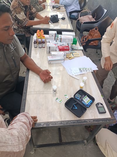 In Cotgadong, Indonesia, an outdoor scene unfolds with individuals seated around a table laden with medical supplies. The table, a light-colored surface with a worn metal frame, is covered with various medications, including boxes of "Fimol Paracetamol" and "AMBROXOL 30 MG," as well as bottles of syrup and blister packs of pills. A glucose meter and its accessories, including lancets and alcohol swabs, are neatly arranged in a black case, suggesting a medical check-up or distribution of supplies. A person in the foreground, wearing a gray vest over a patterned shirt, has their arm resting on the table, their hand open. To their left, another individual in a patterned brown and beige garment is visible. In the background, a person in a white coat and another in a light-colored uniform are present, with chairs, some occupied and some not, surrounding the area. The overall environment suggests a community health event or a mobile clinic operation.