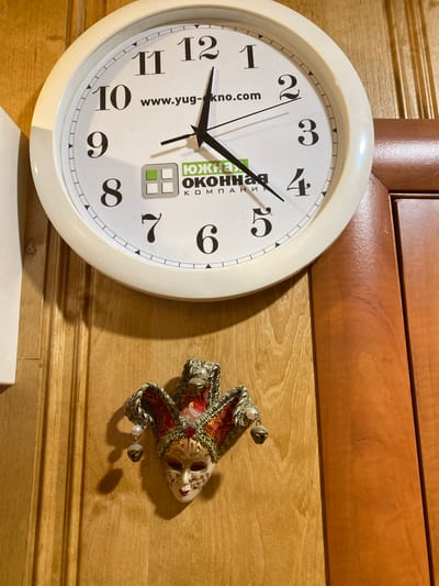 A round, white analog clock with black Arabic numerals hangs on a wooden paneled wall. The clock face features a website address, "www.yug-tkno.com," and a logo that appears to be for a window company. Below the clock, a decorative Venetian mask, designed as a jester's face with red, gold, and white accents, hangs on the wall. The mask has bells and pearls attached to its sides. The lighting in the scene casts a distinct shadow of the mask on the wall. The image does not contain any elements that would place it in Yokohama, Japan.