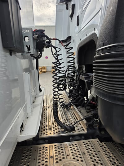 The image shows the underside of a truck, specifically focusing on the connection area between the truck cab and the trailer. The primary subjects are various mechanical components, including coiled cables, air lines, and metal brackets. The scene is set outdoors, likely in a truck yard or loading area, given the presence of pallets in the background and the overall industrial nature of the equipment.
The image captures a static view of these truck components, suggesting no specific activity is taking place at the moment the photo was taken. The lighting indicates it is daytime, with a bright, overcast sky visible in the distance, hinting at potentially cloudy weather. The location is identified as Zeppelinheim, Germany, which provides geographical context for this industrial scene.
Notable details include the prominent coiled black cables, which are likely electrical and air lines for connecting the truck to its trailer. There are also several metal components, some perforated with holes, which appear to be part of the trailer hitch or support structure. A small black rectangular object, possibly an electronic module, is visible in the lower left corner, with the partial text "EC..." on a metal bracket beneath a black box. The overall impression is one of robust, functional machinery.