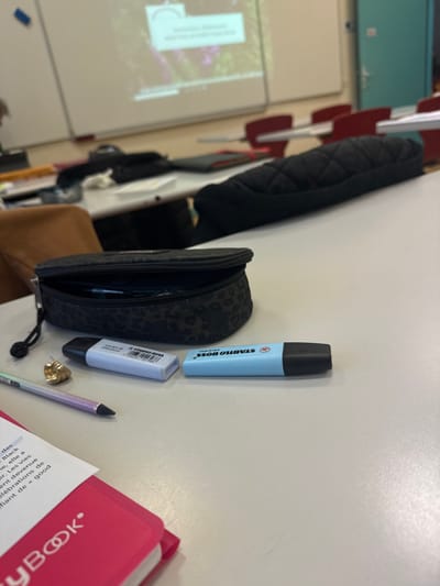 The image captures an interior view of a classroom or lecture hall in Albi, France, focusing on a desk in the foreground. On a light grey laminated desk surface, a partially unzipped black pencil case with a subtle dark leopard print pattern is positioned on the left. Two "STABILO BOSS ORIGINAL" highlighters lie horizontally to its right: one light blue with a black cap and one light purple with a white cap, displaying a barcode. A light purple pen with a black tip and clip rests horizontally to the left of the highlighters. Between the pen and the purple highlighter, a single, irregularly shaped gold-colored earring is present. In the bottom-left corner, a pink "MYBOOK" notebook is visible, with a page open displaying partial French text including "...des... Black ...elle a... Les vies ...devenue célébrations de ...fiant de - good".
In the mid-ground, a second light grey desk holds a black quilted bag or case. Behind this, several chairs with red backs are visible, receding into the background, along with a brown chair. The background features a large white whiteboard mounted on a wall, onto which a digital projector displays an image of foliage. Overlaid on the projected image is a white box containing partial French text, including phrases like "...différentes et leurs évolutions". A light teal door with a silver handle is visible on the far right of the frame. No individuals are clearly identifiable in the scene.