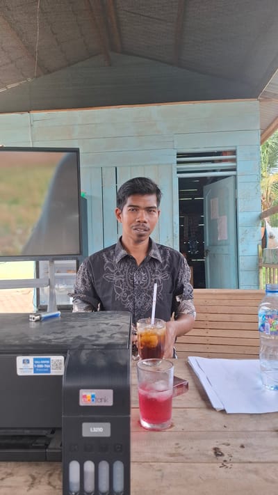 A man with dark hair and a mustache, wearing a black collared shirt with a white patterned design, is seated at a wooden table. He is looking towards the camera. In front of him are two glasses with drinks, one with ice and a straw, and a bottle of water. To his left is a black printer with an "ecotank" logo and a model number "L3210". A screen or monitor is also visible to his left. The background shows a light blue wooden structure, possibly a building or shelter, with an open doorway. Wooden planks form seating or a counter area behind the table. The environment appears to be outdoors, with vegetation visible in the background. The scene is set in Grong-grong, Indonesia.