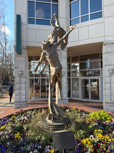 A bronze sculpture depicts a horse-like figure carrying a person playing a violin. The sculpture stands outside a modern building with large windows and a glass-door entrance marked "210." The sculpture is placed among colorful flowers. A plaque sits beside the sculpture, detailing the art piece and its contributors. A person is walking on the sidewalk.