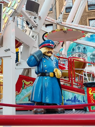 This is a medium shot, eye-level, outdoor photograph of a statue of a man dressed as a historical police officer. The statue is blue with gold accents and appears to be part of a carnival ride, possibly a Ferris wheel given the large white structure in the background. The officer has a handlebar mustache and is saluting with his right hand to his eye. He is wearing a blue uniform with gold buttons, a black belt with a gold buckle, and a blue hat with a red stripe and gold insignia. The background shows the colorful and elaborate machinery of a carnival ride, including lights, metal supports, and painted panels depicting cityscapes. The scene is brightly lit, suggesting it is daytime. Red ropes and barriers are in the foreground, indicating a public viewing area. The overall atmosphere is festive and reminiscent of a funfair. There is a sign in the background with some illegible German text.