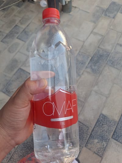An overhead shot shows a hand holding a clear plastic bottle of OMAF Natural Mineral Water with a red label, partially filled with water. The bottle has a red cap. The background consists of a grey tiled surface.