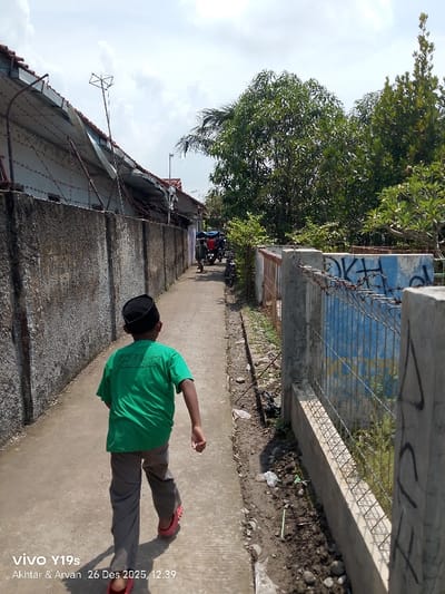 A child in a green shirt, grey pants, and red sandals walks away down a narrow alley. High concrete walls line the left side, topped with barbed wire and fences, while a fence with graffiti and overgrown vegetation marks the right. Further down the alley, figures, vehicles, and lush trees appear. The sky is partly cloudy. The image is stamped "vivo Y19s, Akhtar & Arvan 26 Des 2025, 12.39."