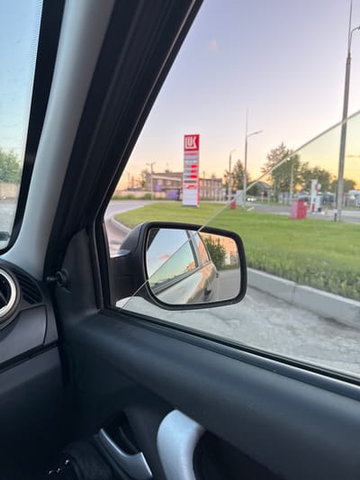 The image is taken from inside a car, looking out the passenger side window. The scene depicts a roadside view with a Lukoil gas station prominently visible in the background. The sky is clear with a soft gradient from blue to orange, suggesting it is either sunrise or sunset. Trees and buildings are scattered along the roadside. In the foreground, the interior of the car is visible, including the door panel, window frame, and side mirror. The side mirror reflects a portion of the car's interior and the outside environment. The overall impression is a snapshot of a journey or a stop at a gas station.
