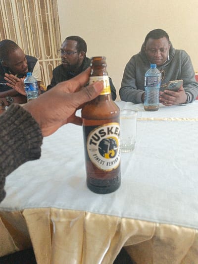 Three men sit around a table. Two men talk and one looks at his phone. Water bottles and a glass are on the table. A hand holds up a Tusker beer bottle, obscuring part of the scene. The table is covered with a white tablecloth trimmed with gold. Vertical blinds appear in the background.