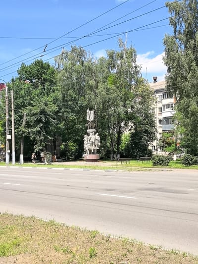 The image depicts a street scene in Bryansk, Russia, featuring a monumental sculpture on the right side of the frame. The sculpture, made of stone, is a tall structure topped with what appears to be draped figures and includes a lower section with several standing figures in a group. The sculpture is situated on a circular base and is surrounded by greenery.
In the background, a multi-story apartment building with balconies and windows is visible, suggesting a residential area. Trees are abundant throughout the scene, with lush green foliage framing the buildings and the sculpture. Overhead, power lines are strung across the sky, indicating urban infrastructure.
In the foreground, a paved road with white lane markings is prominent, suggesting a thoroughfare. A grassy verge with some dry patches runs alongside the road. On the left side of the street, a pedestrian is walking away from the camera, and further left, a utility pole with electrical equipment is present. Two park benches are visible on the grass area near the sculpture, implying a public space for rest. The sky is blue with scattered white clouds.