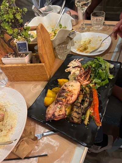 The scene is a table at a restaurant in Limenaria, Greece (latitude: 40.628, longitude: 24.575). A plate of food is in the center of the table, containing grilled squid, eggplant, peppers, and potatoes. There are also plates of food, a glass of wine, and other miscellaneous items on the table. The person in the background is partially visible, wearing jeans. The environment is a casual restaurant setting.