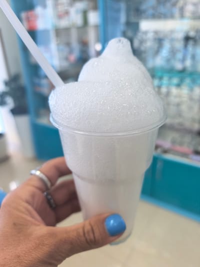 In this close-up shot, a hand with blue-painted fingernails and several rings, including a silver band and a darker, possibly beaded ring, holds a clear plastic cup filled with a frothy, white beverage. A white straw extends from the foamy top of the drink. The beverage appears to be a soda or a milkshake with a significant amount of foam piled high above the rim of the cup. The background is blurred but suggests an indoor setting with light-colored flooring and shelves displaying items, possibly in a store or establishment. The dominant colors in the background are light blue and white, with some hints of red and other muted colors on the shelves. The lighting suggests it could be daytime. There is no visible text in the image. The focus is on the drink and the hand holding it, creating a sense of a personal moment enjoying a beverage.