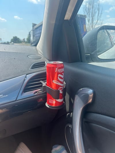 The image shows the interior of a car, focusing on a red can of Freeway Cola placed in a black cup holder. The cup holder is attached to the car's dashboard, positioned to the right of the air vents. The dashboard is dark gray and textured, with a silver trim piece to the left of the air vents that has the word "Tiline" visible. The can itself is mostly red with white lettering. At the bottom of the can, a Nutri-Score label indicates a rating of A, B, C, D, and E. A black handle for the car door is visible to the right of the cup holder. In the background, through the car's windows, an outdoor scene is visible, suggesting the car is on the road. The sky is blue with some clouds. The crops show parts of the car's interior, the exterior environment, and close-ups of the can and cup holder. The location is identified as Tsaratsovo, Bulgaria.
