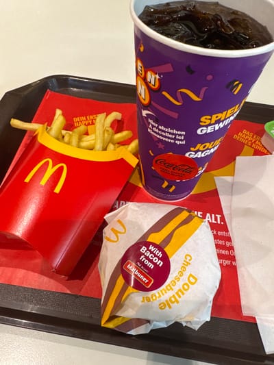 The image displays a McDonald's meal served on a black tray. The meal includes a large purple cup filled with a dark liquid, likely Coca-Cola Zero, with German and French text on the cup. There is also a box of McDonald's fries. Additionally, there is a "Double Cheeseburger with Bacon from Malbuner". The food is sitting on a red paper liner with German text and McDonald's logos. Based on the text "DEIN ERSTE HAPPY MEAL DEINES KINDES" and the branding in German, the location is likely a McDonald's in a German-speaking region of Switzerland. Due to the presence of Malbuner bacon (from Switzerland), it is more specifically likely to be a McDonald's in Switzerland, and most likely in the city of Spreitenbach, Switzerland.