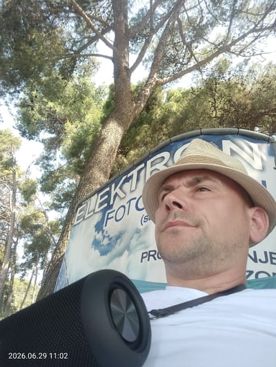 A man wearing a straw hat takes a selfie outdoors. He is positioned in the foreground, with the lower part of his body and a black speaker to his left. Behind him, a large banner displays text that includes "ELEKTRON FOTO" and other partial words in a blue and white cloud-filled sky design. The background also features the trunk and branches of a pine tree, with more trees visible in the distance. The lighting suggests it is daytime, possibly a sunny day, casting shadows and highlighting the man's face as he looks upward and to the right. The man appears relaxed or contemplative. A timestamp "2026.06.29 11:02" is visible in the bottom left corner of the image. The scene appears to be set in a warm, possibly Mediterranean, climate, consistent with the context of Krk, Croatia.
