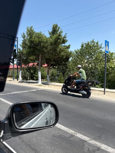 A person wearing a green shirt, denim shorts, and a white baseball cap is riding a black electric scooter on a paved road. The scooter has the word "CHEETAH" on its side. The road has a white painted line down the center. To the left of the road, a traffic light is visible, displaying a green signal. A pedestrian crossing sign is posted to the right of the road. Trees line the side of the road, and some have their lower trunks painted white. In the foreground, a car's side mirror reflects a portion of the car's interior and the road. The sky is clear and blue. The scene appears to be a street in the city of Kahramon, Uzbekistan.