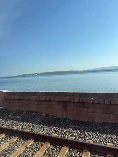 The image is a landscape photograph taken outdoors, oriented vertically. The scene depicts a body of water adjacent to a railway track.
In the foreground, on the right side of the frame, are railway tracks. These consist of two parallel metal rails laid on wooden sleepers, embedded in a bed of dark gray ballast (small stones). To the left of the tracks, a low wall constructed of reddish-brown bricks runs horizontally, creating a distinct border.
The middle ground is dominated by a wide expanse of blue water, likely a sea or a large lake, stretching horizontally across the frame. The surface of the water is slightly rippled, indicating a gentle breeze. Along the distant horizon, a faint outline of land can be seen, with mountains or hills rising in the background. Some man-made structures, possibly industrial buildings or a town, are visible on the shoreline in the far distance.
The sky occupies the upper left portion of the image. It is a clear, bright blue, with a gradient becoming lighter towards the horizon. A thin white contrail from an aircraft is visible high in the sky.
The overall impression is one of a bright, clear day. The sunlight suggests it is daytime, likely late morning or early afternoon given the brightness and absence of long shadows. The scene evokes a sense of peacefulness and openness, with the juxtaposition of the man-made railway and wall against the natural elements of water, land, and sky. No people or specific activities are visible. There is no visible text in the image. The location context provided is Bages, France, which aligns with a coastal or lakeside setting.