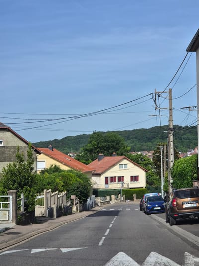 The image depicts a street scene in Audun-le-Tiche, France. The view looks down a residential street with a slight incline. On the right side of the street, two parked vehicles are visible: a brown Dacia Duster and a blue car. The street is paved, with a white dashed center line and a pedestrian crossing indicated by white triangular markings.
On the left side of the street, several houses are present, varying in architectural style and color. The houses are surrounded by greenery, including trees and bushes. Overhead, power lines stretch across the sky, supported by a concrete utility pole. In the background, a hillside covered with dense forest rises, suggesting the town is situated near a forested area. The sky is clear and blue, indicating a sunny day. A small cemetery or memorial area with numerous stone structures is located adjacent to the road in front of the yellow house.