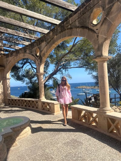 The image depicts an outdoor scene within the grounds of the Marivent Palace in Cas Català-ses Illetes, Spain. A single female subject is positioned centrally, walking forward on a light-colored, pebbled stone path. She has long, light-colored hair, wears dark sunglasses, a pink long-sleeved, tiered dress, and dark flip-flops, with a black backpack visible on her shoulders.
Architectural elements dominate the foreground and midground. A series of light-colored stone arches with decorative circular openings are visible overhead and to the right, supporting a wooden pergola structure. A matching stone balustrade, featuring X-shaped patterns between its supports, runs parallel to the path on the right. To the left, a multi-tiered stone water feature or fountain is partially visible.
Beyond the architectural structures, a clear blue body of water, likely the Mediterranean Sea, is visible, populated by numerous white sailboats and yachts. Green pine trees and other vegetation line the coastal hillside. A small, vegetated island or rocky outcrop is present in the bay. The environment is under bright, clear sunlight, indicated by distinct shadows cast by the arches, balustrade, and the subject onto the path. The sky is uniformly blue with no cloud cover.