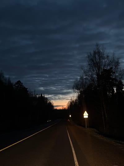 The image depicts a road at dusk, with a dramatic sky filled with striated clouds. On the right side of the frame is a paved road with a solid white line marking the edge. A yellow stripe is also present on the road’s edge. A road sign is visible, illuminated by a lamp, indicating a curve in the road. Trees line the roadside, silhouetted against the dim sky. The location may be Izvestkovyy, Russia, based on the prompt's request, though this cannot be definitively confirmed from the visual information alone. There are no people visible in the scene.