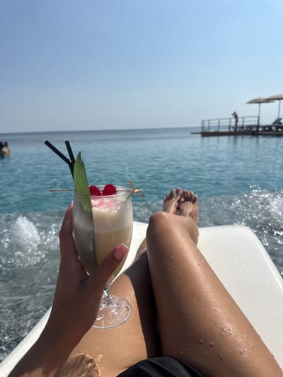 The image presents a first-person view from a white poolside lounge chair, focusing on a person's hand holding a cocktail and their legs. In the lower left, a hand with a French manicure holds a stemmed glass filled with a light-colored, frothy cocktail, garnished with two red cherries, two black straws, a green leaf, and a wooden pick. The person's tanned legs, speckled with water droplets, extend into the lower right, resting on the white lounge chair. Turbulent water, possibly from jets, is visible in the pool to the left of the hand. The midground features a serene blue infinity pool, visually merging with a large, calm body of water under a clear blue sky. In the distant background, a dock-like structure with railings extends over the water, where a silhouetted person stands, and two large sun umbrellas are present. A blurred individual is partially submerged in the water on the far left. The scene is situated in Hlibivka, Ukraine.