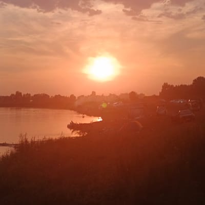 The image depicts a sunset over a body of water, likely a river or lake, adjacent to a shoreline. The sky is filled with orange and pink hues, with some clouds scattered across it. The sun is a bright, glowing orb near the horizon. On the shore, there are several tents and vehicles, suggesting a campsite or gathering. Silhouettes of trees and buildings are visible in the background, hinting at a nearby settlement or natural landscape. The water reflects the colors of the sky, creating a serene atmosphere. The overall impression is one of a tranquil evening scene by the water.
