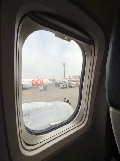 The image was taken from inside an airplane window, looking out at an airport tarmac. The exterior of the aircraft's wing is visible in the foreground, with the window frame of the airplane interior surrounding the view.

In the background, a GOL Airlines plane is parked, identifiable by the prominent "GOL" logo on its fuselage. Other aircraft and airport vehicles are also visible on the tarmac. A single individual wearing a yellow vest is seated on the ground near the aircraft. The sky appears overcast.

This scene is characteristic of the operations at São Paulo/Guarulhos International Airport (GRU) in Guarulhos, Brazil, a major hub for GOL Airlines.