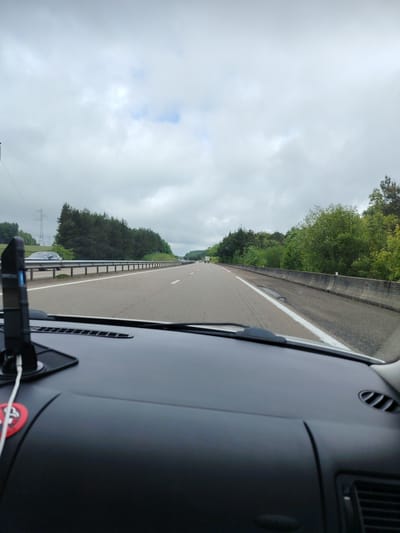The image shows a view from the dashboard of a car driving on a highway near La Pellerine, France. The highway is mostly empty with light traffic visible in the distance. There are trees alongside the road. The sky is overcast. A car mount with a phone is attached to the dashboard. A sticker with a heart symbol is also visible on the dashboard.