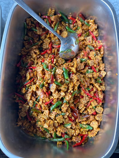 A top-down, slightly oblique view captures a rectangular, stainless steel serving tray filled with a textured Indonesian dish in Kutalhoksukon, Indonesia. The dish consists primarily of small, irregularly shaped, golden-brown pieces, consistent with fried tempeh, mixed with vibrant red and green chili peppers (likely birds-eye and larger green chilies) and possibly finely sliced aromatics. A metallic silver serving spoon or ladle is partially submerged in the food, extending diagonally from the upper left towards the lower right of the tray, indicating readiness for serving. The tray exhibits minor scuffs and reflections on its metallic surface. No individuals are visible. The immediate background is out of focus, suggesting a food service environment.