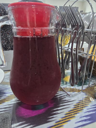 A close-up, eye-level shot features a large, clear glass pitcher filled with a dark red liquid, likely juice or a beverage. The pitcher has a red lid and is positioned on a table with a colorful, patterned tablecloth. To the right of the pitcher, a metal rack holds several sets of silverware, primarily forks, with some spoons visible. In the background, a person wearing a dark, floral-patterned garment is partially visible on the left. The setting appears to be indoors, possibly a dining area, with some elements of the environment, like blinds and a ventilation unit, visible in the background. The overall impression is a still-life composition with a focus on the beverage and tableware.
