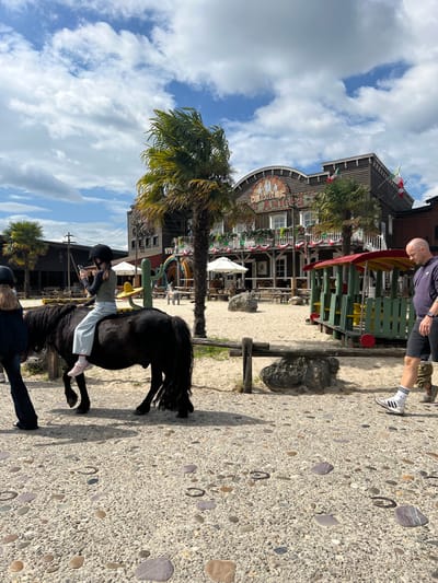 The image depicts an outdoor scene in Collendoorn, Netherlands, likely a theme park or entertainment venue, given the rustic Western-style architecture of the "Hotel Los Amigos" building in the background. In the foreground, a young person wearing a helmet, dark long-sleeved shirt, and light blue jeans is riding a small black pony. The rider is holding a smartphone in their left hand. To the left of the pony, another person, mostly out of frame, is walking. On the right, a bald man wearing a dark hoodie and black shorts with white socks and sneakers is walking away from the camera. The ground is a mix of sand and textured pavement with visible pebbles. Behind the pony, a wooden fence separates the sandy area from the paved walkway. The background features palm trees, white umbrellas, tables and chairs, and a colorful playground structure resembling a train. The sky is partly cloudy.
