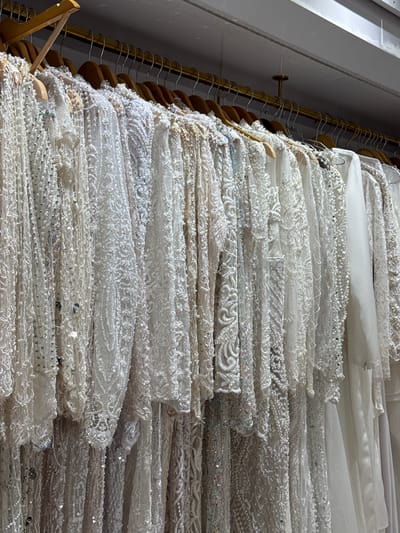 A row of ornate white dresses, likely wedding or formal gowns, are displayed on hangers from a brass rod in what appears to be a retail setting. The dresses feature intricate beadwork, sequins, and lace detailing. The garments are densely packed, creating a textured and shimmering visual effect. The background is out of focus, showing a ceiling and possibly other clothing racks. The overall impression is one of a boutique or bridal shop.