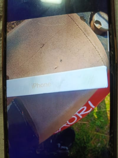 The image displays a close-up, angled shot of an iPhone box resting on a textured, light brown surface. The word "iPhone" is visible in a light, almost transparent font on the box. The background is a blur of green grass and red lettering that appears to be part of a logo or text, partially obscured. The scene is indoors, likely on a table or floor given the lighting and the presence of a textured surface and grass. There is no information about the city of Svoboda, Russia, or any individuals present.